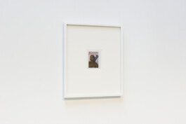 A photographic art work by Jeanetta Rich at The Bag Los Angeles in 2023. The Polaroid photograph is a self portrait of Jeanetta Rich. The artist holds a metal gag in her mouth and holds a purple cloth to her chest while looking off screen. The art work is a Polaroid photograph in a large white frame. The work hangs on a white wall of the gallery. Ezequiel Olvera